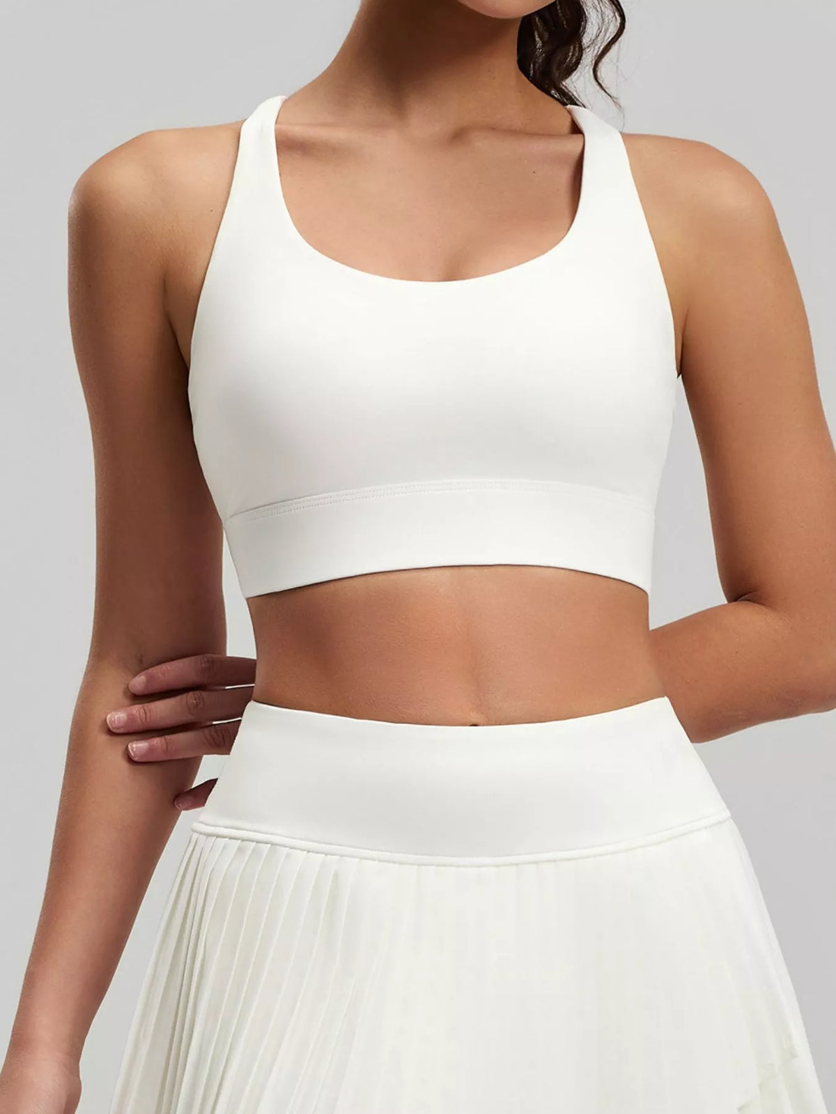 Model wearing white active bra and pleated skirt with one hand resting across the waist